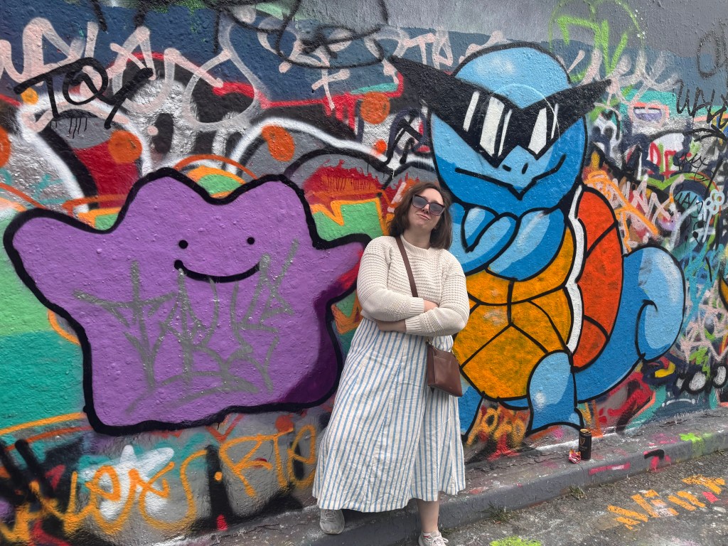 Marisa stands in front of a graffitied wall, featuring the Pokemon Ditto and Squirtle. The Squirtle is the iconic sunglasses squirtle with arms crossed; Marisa imitates this pose with her own sunglasses and crossed arms.